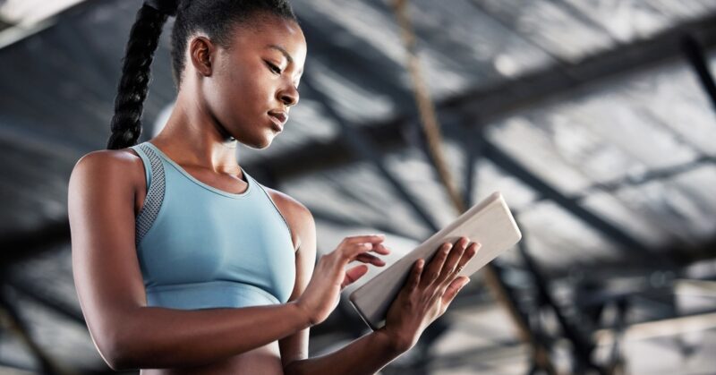 A low-angle view of a young Black woman in a light-blue athletic outfit using a tablet at a facility.