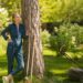 A woman wearing denim overalls leans against a tree holding a rake beside a lawn mower in a sunny green backyard.