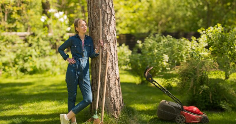 A woman wearing denim overalls leans against a tree holding a rake beside a lawn mower in a sunny green backyard.