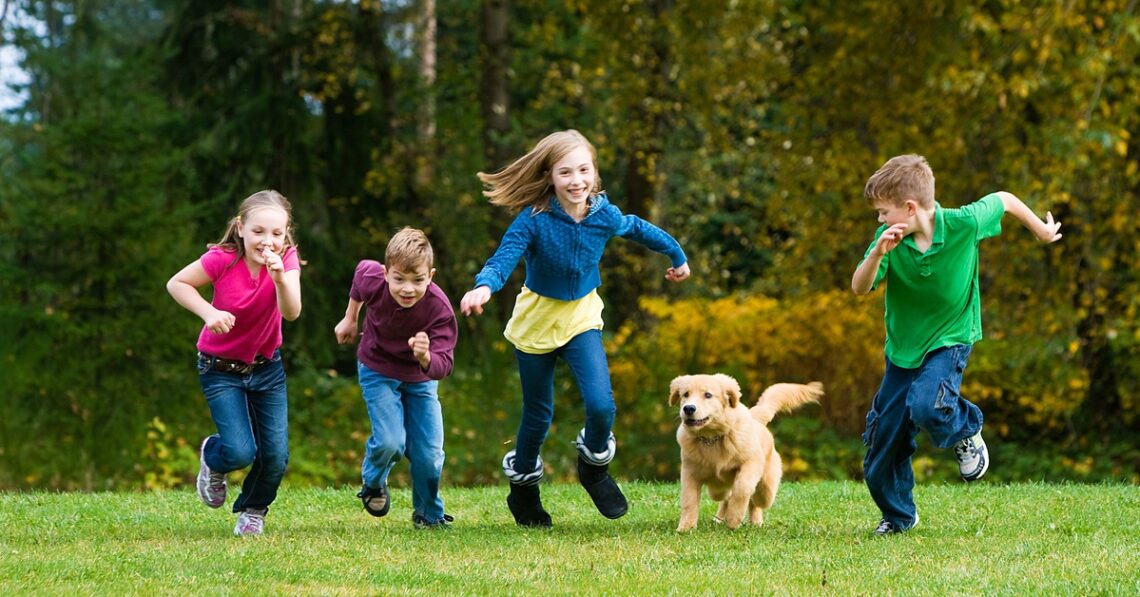 Four kids playing in the backyard, running and smiling with a Golden Retriever puppy. The grass is bright green.
