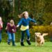 Four kids playing in the backyard, running and smiling with a Golden Retriever puppy. The grass is bright green.