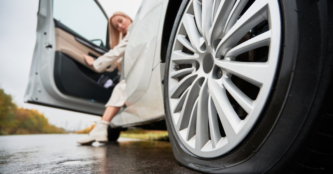 A close-up of a flat tire on a white car. A woman opens the driver's side door and has one foot on the ground.