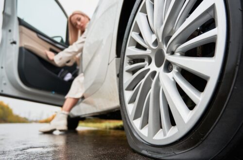 A close-up of a flat tire on a white car. A woman opens the driver's side door and has one foot on the ground.