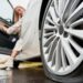 A close-up of a flat tire on a white car. A woman opens the driver's side door and has one foot on the ground.