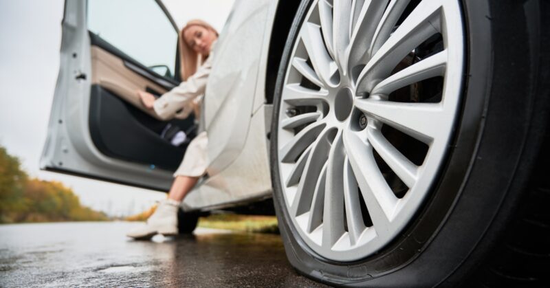 A close-up of a flat tire on a white car. A woman opens the driver's side door and has one foot on the ground.