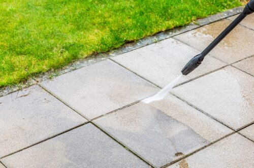 A person operating a pressure washer with a black nozzle pointed at a stone paver patio, spraying the stones.
