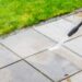 A person operating a pressure washer with a black nozzle pointed at a stone paver patio, spraying the stones.