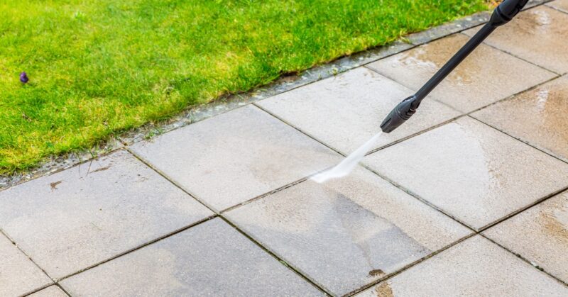 A person operating a pressure washer with a black nozzle pointed at a stone paver patio, spraying the stones.