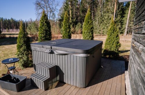 Grey hot tub with a covered lid on a wooden deck, with plastic stairs, a small round table, and a black tub nearby in a backyard.