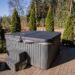 Grey hot tub with a covered lid on a wooden deck, with plastic stairs, a small round table, and a black tub nearby in a backyard.