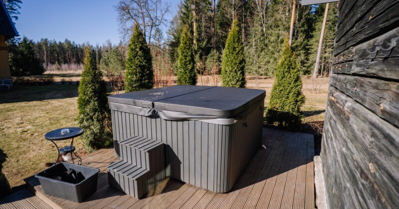 Grey hot tub with a covered lid on a wooden deck, with plastic stairs, a small round table, and a black tub nearby in a backyard.