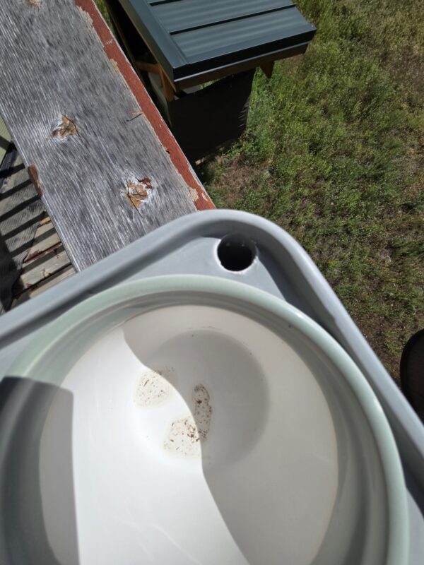 Top-down view of a white bucket with a circular hole in its rim, placed on a weathered wooden beam near green grass.