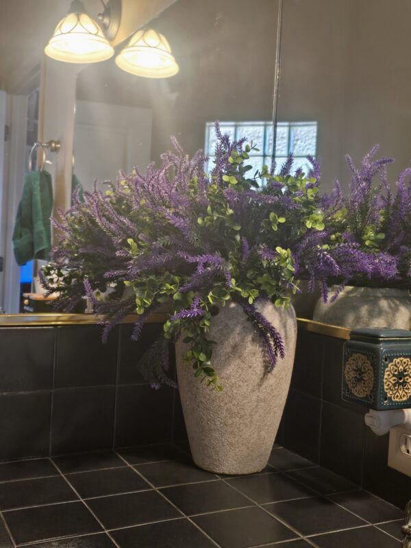 Large stone planter with purple lavender and green foliage sits on a black tiled bathroom counter, reflected in a mirror with warm light fixtures above. Vintage Ceramic Farmhouse Vase with Flowers. flower vase