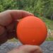 Close-up of a bright orange circular disk held between fingers outdoors, with a forest and hills in the background.