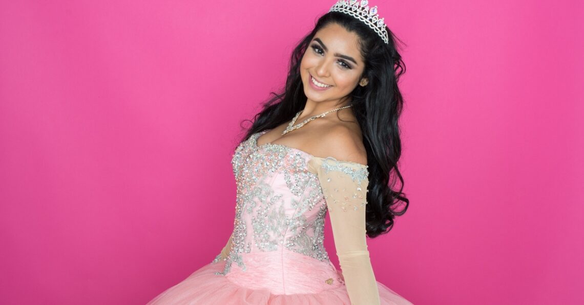 A smiling woman wearing a tiara and pink embellished dress while standing before a solid pink background.