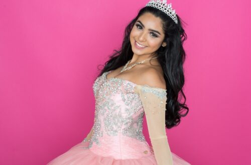 A smiling woman wearing a tiara and pink embellished dress while standing before a solid pink background.