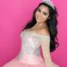 A smiling woman wearing a tiara and pink embellished dress while standing before a solid pink background.