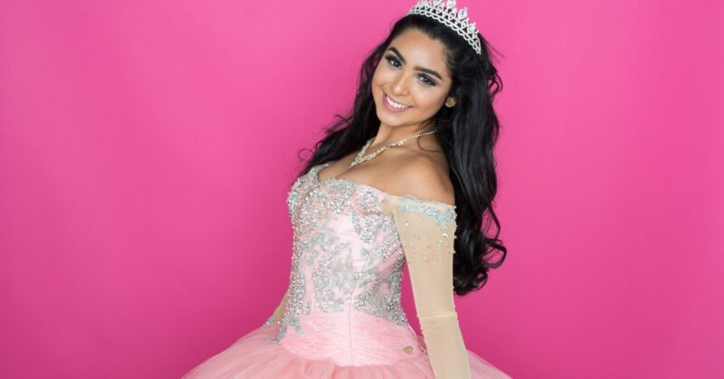 A smiling woman wearing a tiara and pink embellished dress while standing before a solid pink background.