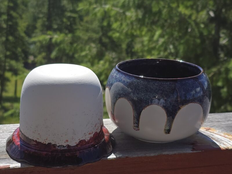 A ceramic pot sit on a wooden surface outdoors, one light gray with a red base and the other dark blue with a white pattern, with green trees in the background.