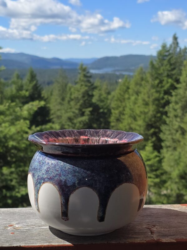 Colorful ceramic pot with blue and purple glaze sits on a weathered wooden railing, with a blurred forest and distant mountains in the background.
