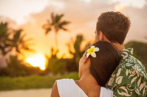 A woman with a white flower in her hair rests her head on a man's shoulder while they watch the sunset on the beach.