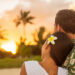 A woman with a white flower in her hair rests her head on a man's shoulder while they watch the sunset on the beach.