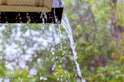 Water pours from the end and underside of a roof gutter during heavy rain. The background shows blurred green trees.