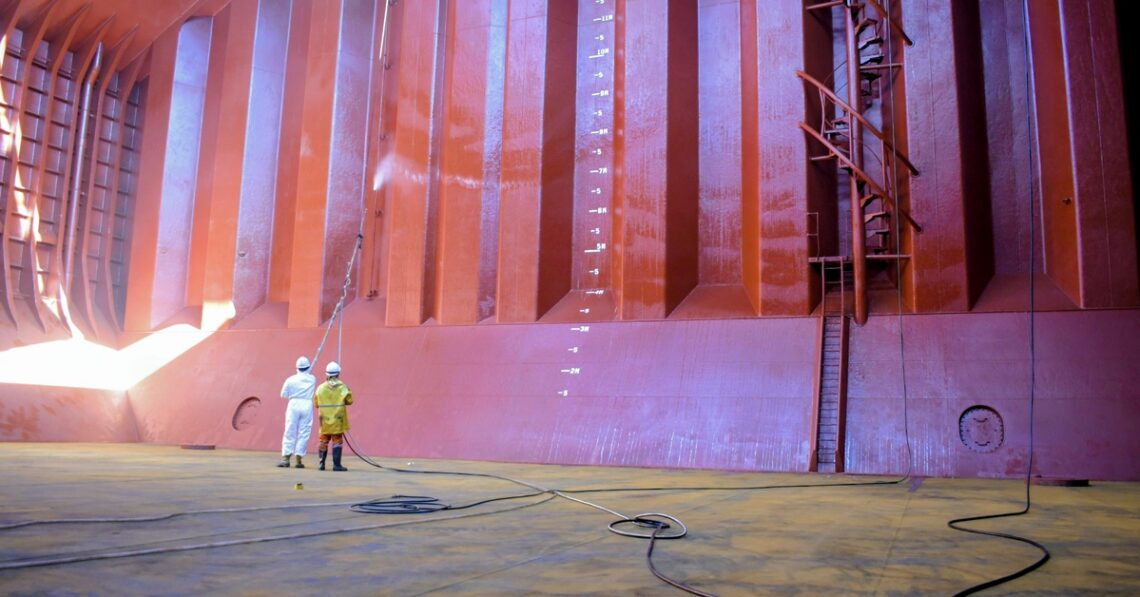 Two workers in protective suits stand with hoses inside a massive pink-orange industrial chamber or ship hull section, inspecting equipment.