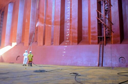 Two workers in protective suits stand with hoses inside a massive pink-orange industrial chamber or ship hull section, inspecting equipment.