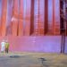 Two workers in protective suits stand with hoses inside a massive pink-orange industrial chamber or ship hull section, inspecting equipment.