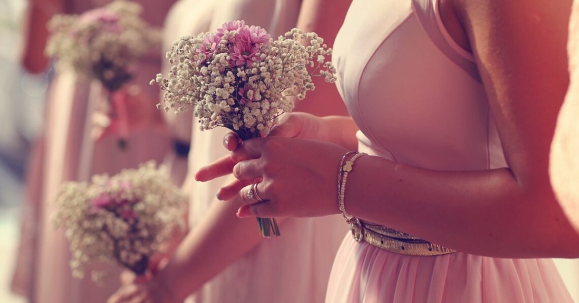 A woman in a pink dress holding a white baby’s breath bouquet tied with a pink ribbon in bright sunlight.