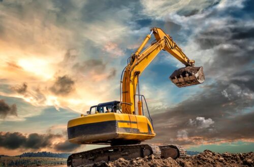 A yellow crawler excavator on dirt tracks lifts a bucket of soil above an earthwork area under a cloudy sunset sky.