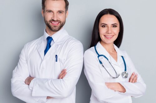 Two medical professionals in white coats stand with arms crossed, smiling at the camera against a plain gray background.
