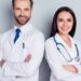 Two medical professionals in white coats stand with arms crossed, smiling at the camera against a plain gray background.