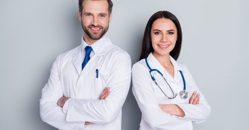 Two medical professionals in white coats stand with arms crossed, smiling at the camera against a plain gray background.
