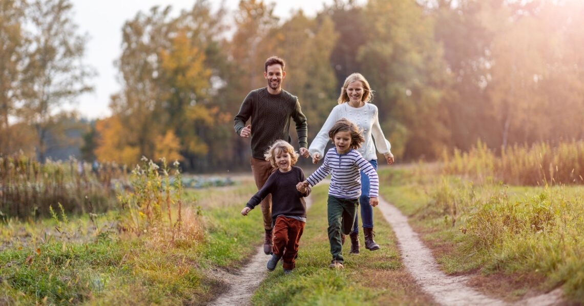 A young family of four running together between dirt tracks on the grass. The two children are holding hands.