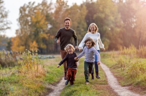A young family of four running together between dirt tracks on the grass. The two children are holding hands.