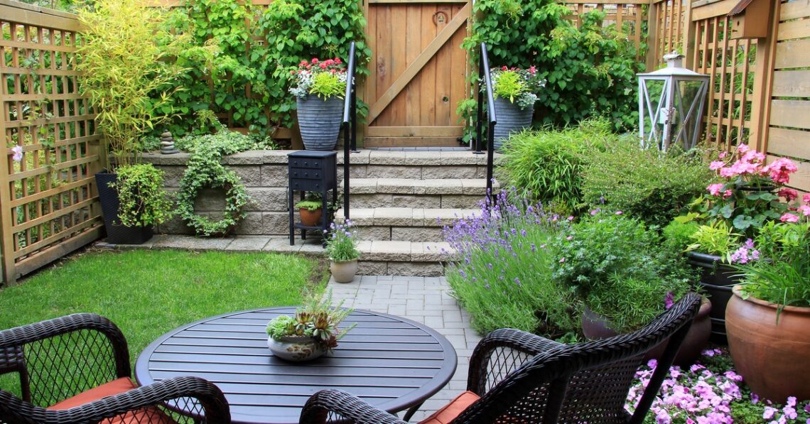 A small fenced-in backyard with various plants, steps leading to a gate, and a small table with two matching chairs.
