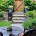 A small fenced-in backyard with various plants, steps leading to a gate, and a small table with two matching chairs.