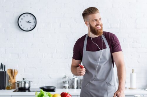 A smiling bearded man in a gray apron wears earphones while cooking with fresh vegetables in a bright modern kitchen.