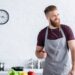 A smiling bearded man in a gray apron wears earphones while cooking with fresh vegetables in a bright modern kitchen.