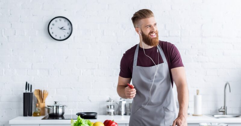 A smiling bearded man in a gray apron wears earphones while cooking with fresh vegetables in a bright modern kitchen.