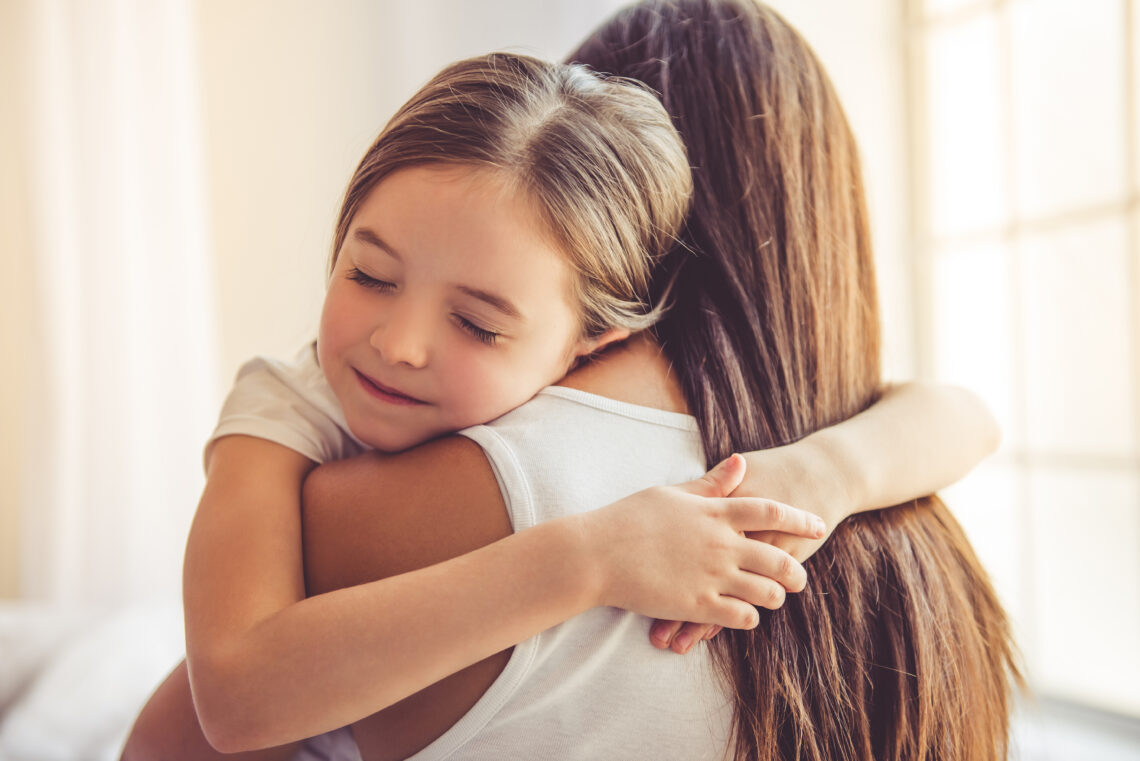 A child with closed eyes hugging a woman in a white shirt near bright windows in a softly lit room together.