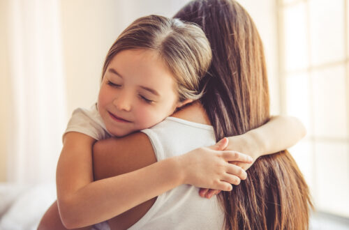A child with closed eyes hugging a woman in a white shirt near bright windows in a softly lit room together.