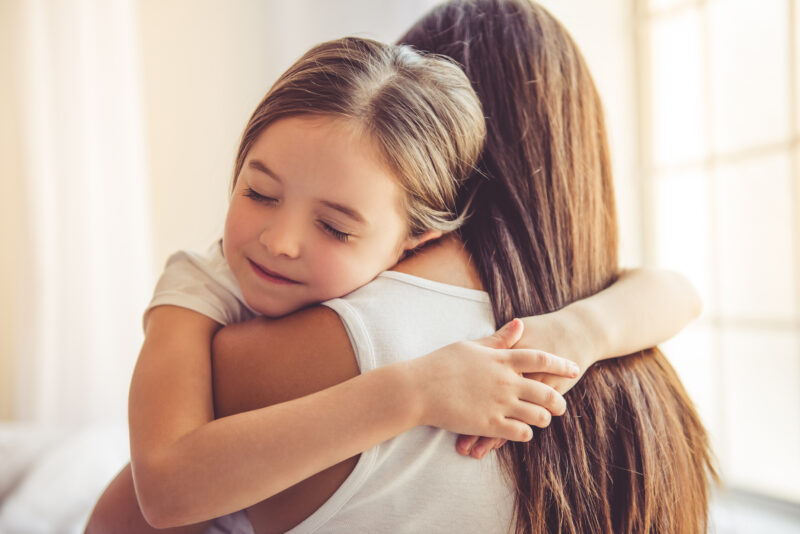 A child with closed eyes hugging a woman in a white shirt near bright windows in a softly lit room together.