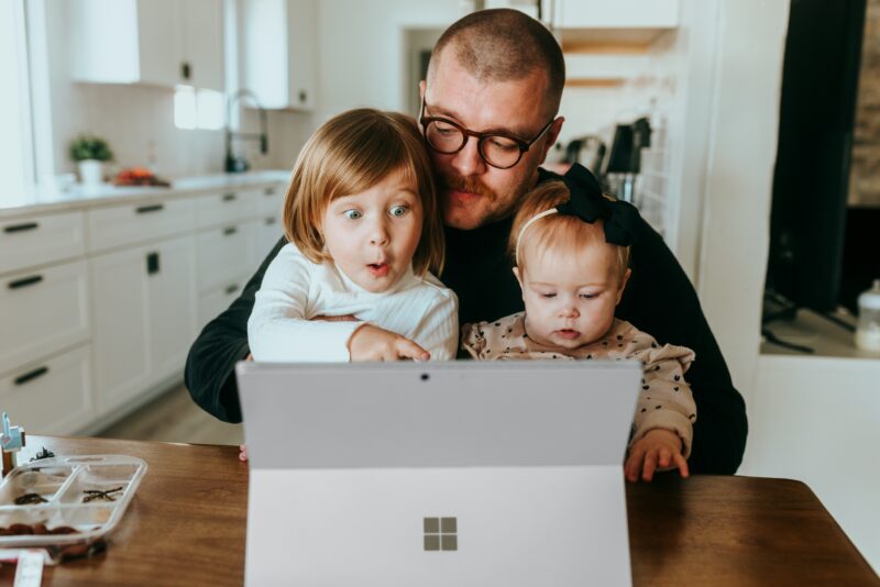 Father with glasses holding two young children as they look at a laptop screen in a bright kitchen.