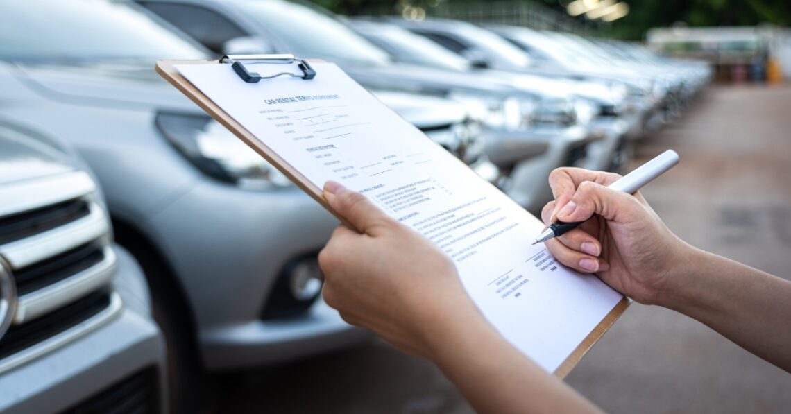 A hand signing a document on a clipboard, with blurred cars parked in rows outside a building behind it.