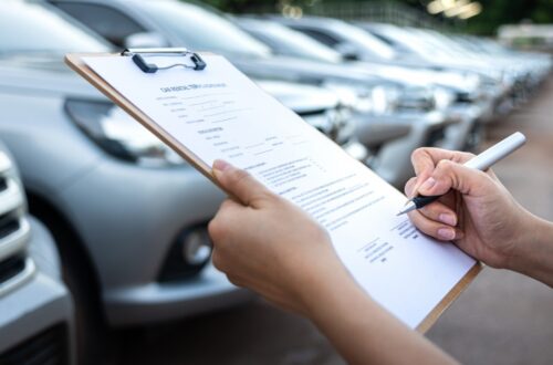A hand signing a document on a clipboard, with blurred cars parked in rows outside a building behind it.