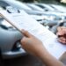 A hand signing a document on a clipboard, with blurred cars parked in rows outside a building behind it.