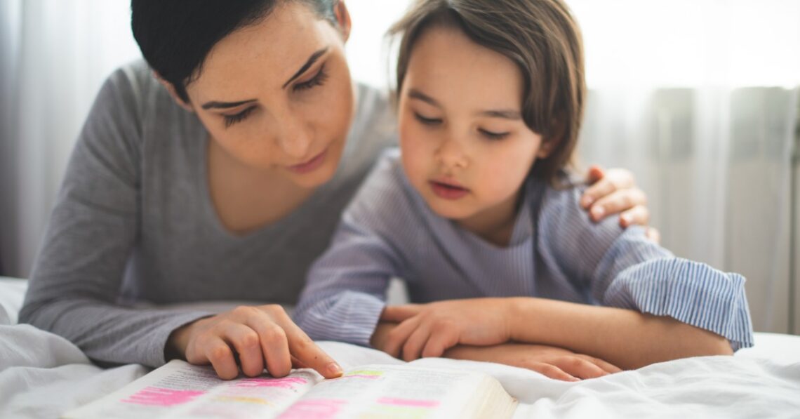 A woman and child kneeling beside a bed while reading an open book together in a bright bedroom.